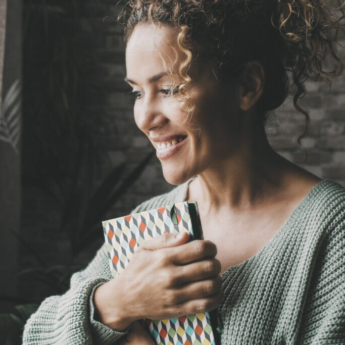 Happy smiling expression woman hold book and look outside the window at home. Dark background. Joyful face portrait of female people in indoor leisure activity alone. Attractive middle age lady indoor